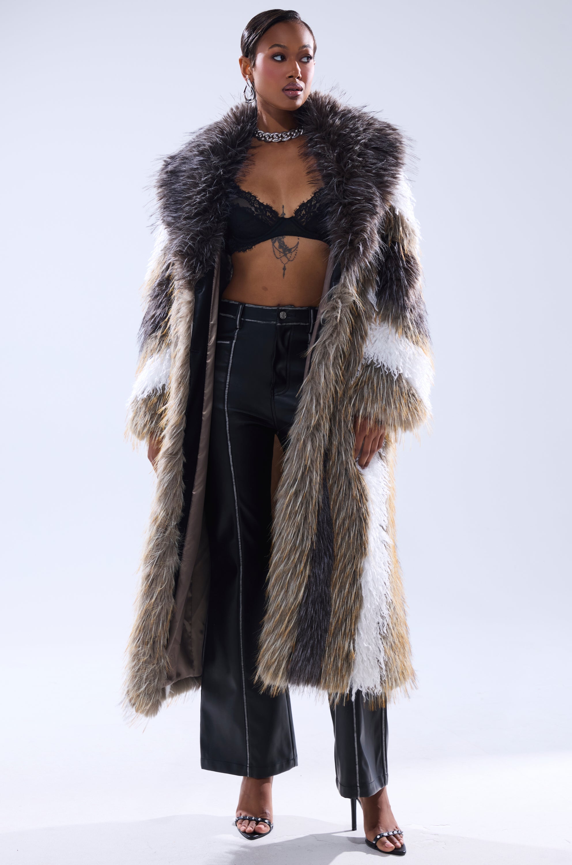 A woman stands confidently against a plain background wearing the POISON IVY FAUX FUR COAT in brown, white, and black, paired with black leather pants with white stitching, a black lace bralette, high heels, and a chunky chain necklace.