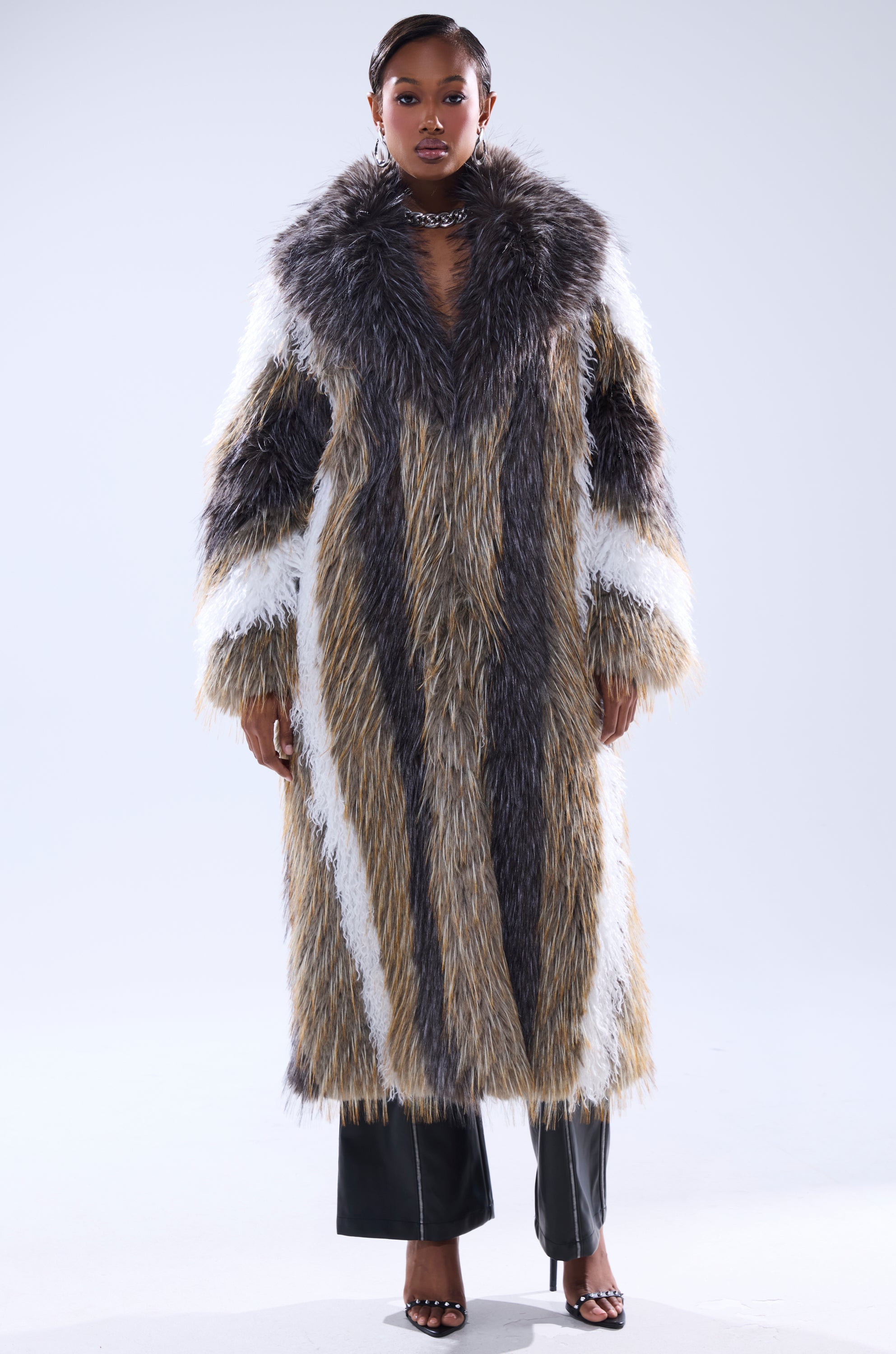 A woman stands facing forward in the POISON IVY FAUX FUR COAT—long and plush with gray, white, and tan vertical stripes—paired with black pants, open-toe heels, and a metallic choker against a plain light background.