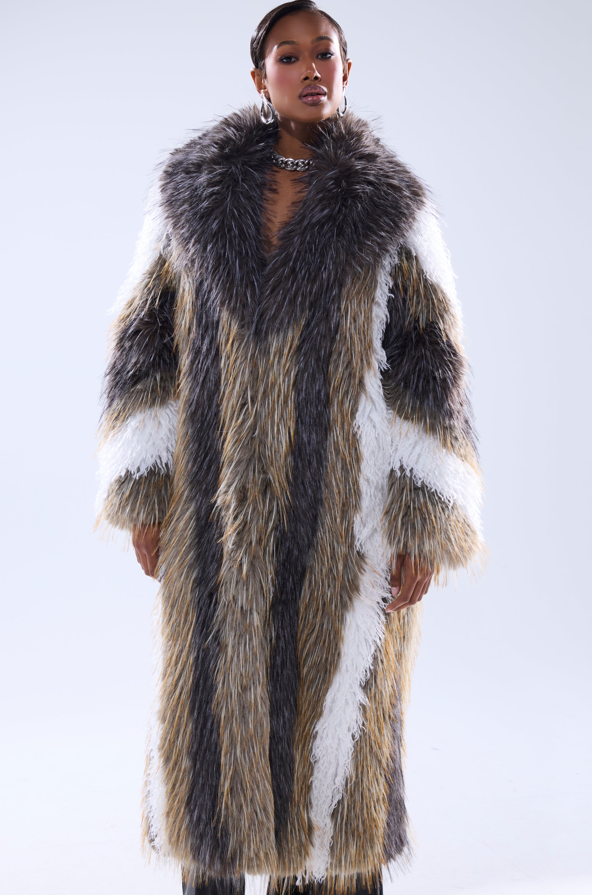 A woman stands facing forward, wearing the POISON IVY FAUX FUR COAT—a long, luxurious piece with large fluffy collars and vertical stripes in brown, gray, white, and tan—against a plain light background.