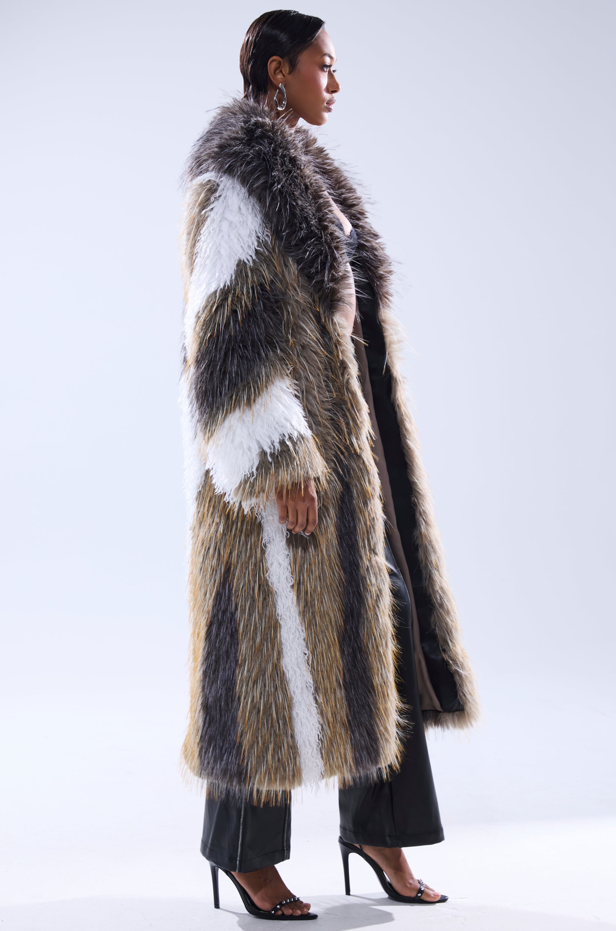 A person in profile models the POISON IVY FAUX FUR COAT, a long, shaggy piece with brown, white, and gray stripes, paired with black pants and high-heeled sandals against a plain light background.