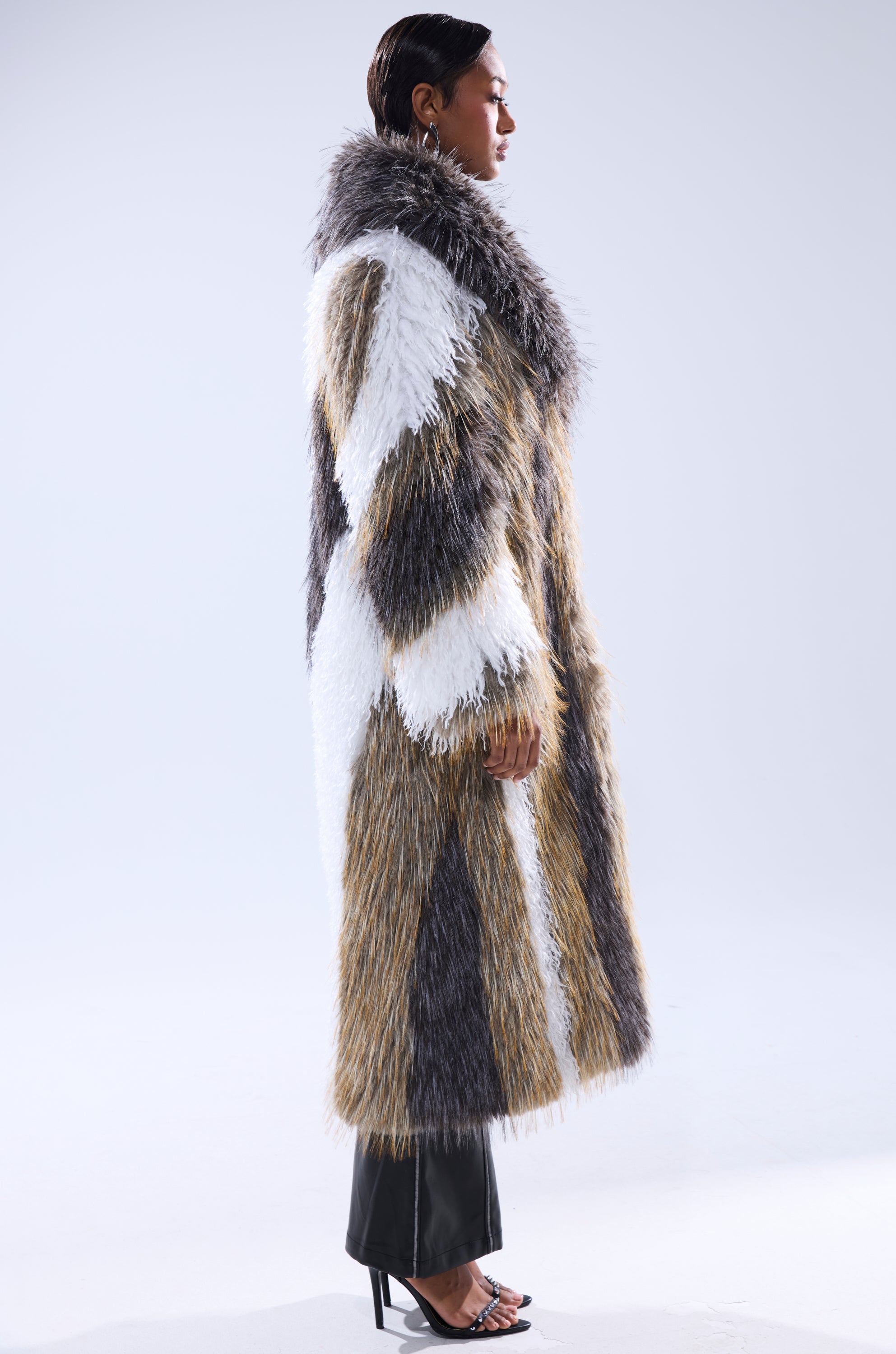 A person in profile wears the POISON IVY FAUX FUR COAT with white, brown, and gray sections, paired with black pants and heeled sandals, standing against a plain light background.