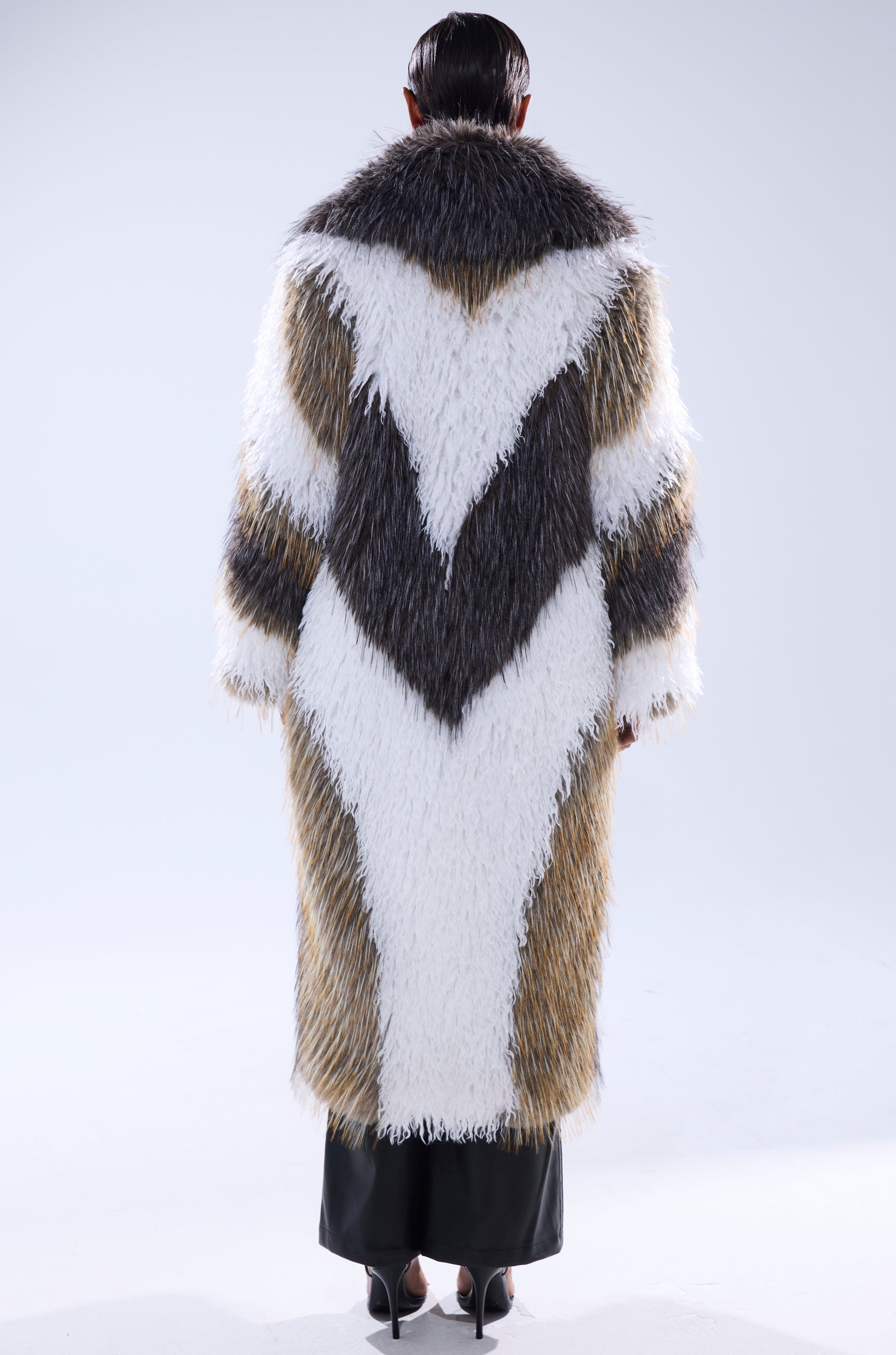 A person faces away, wearing the POISON IVY FAUX FUR COAT—a long coat with a wide collar and bold zigzag patterns in white, black, brown, and gray. The textured faux fur covers them almost to the floor.