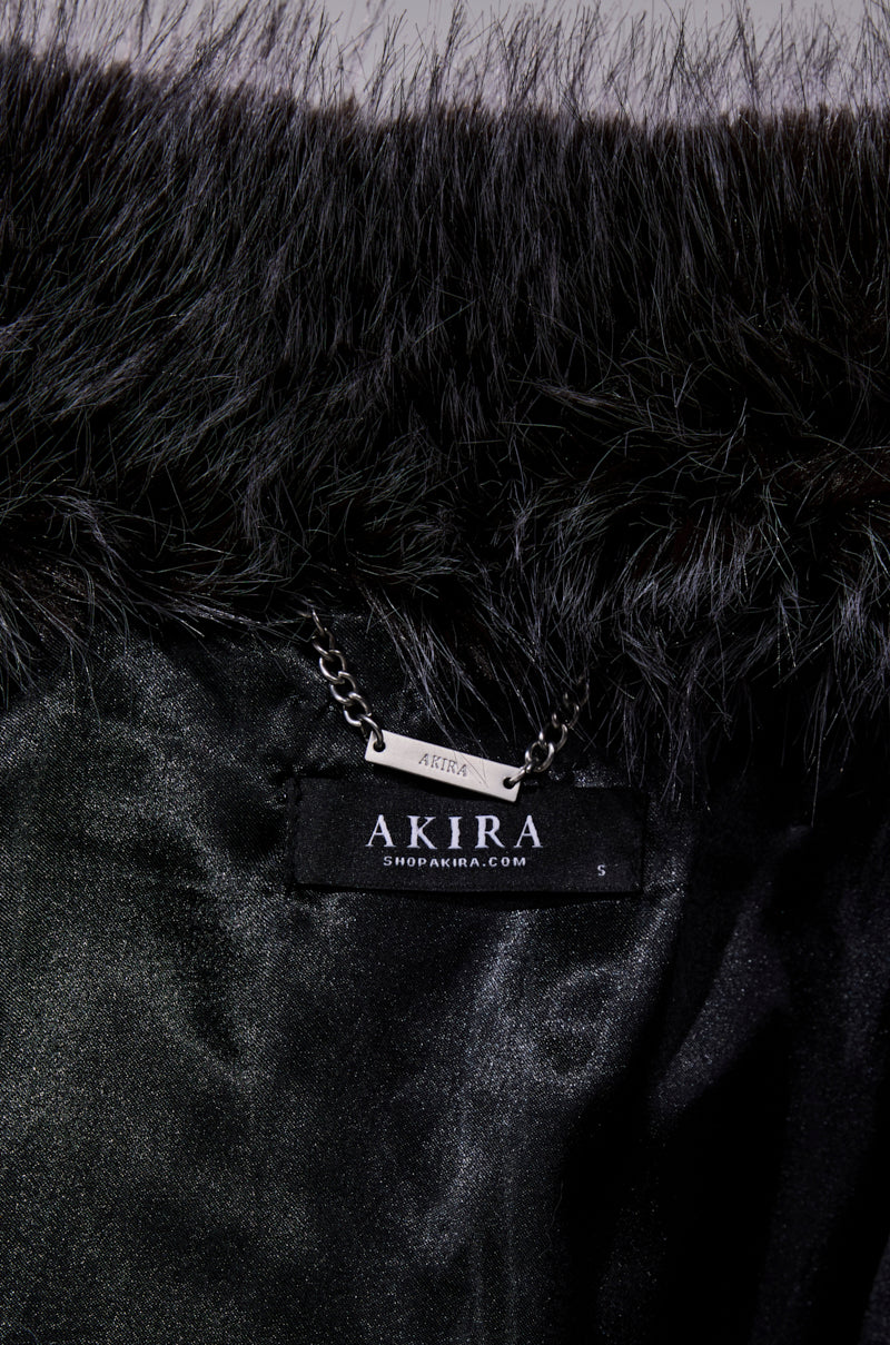 Close-up of the WANT IT ALL FAUX FUR coat, featuring a shiny black faux fur fabric, visible chain detail, and a black label reading "AKIRA" and "SHOPAKIRA.COM." The luxurious material highlights its stylish longline silhouette.