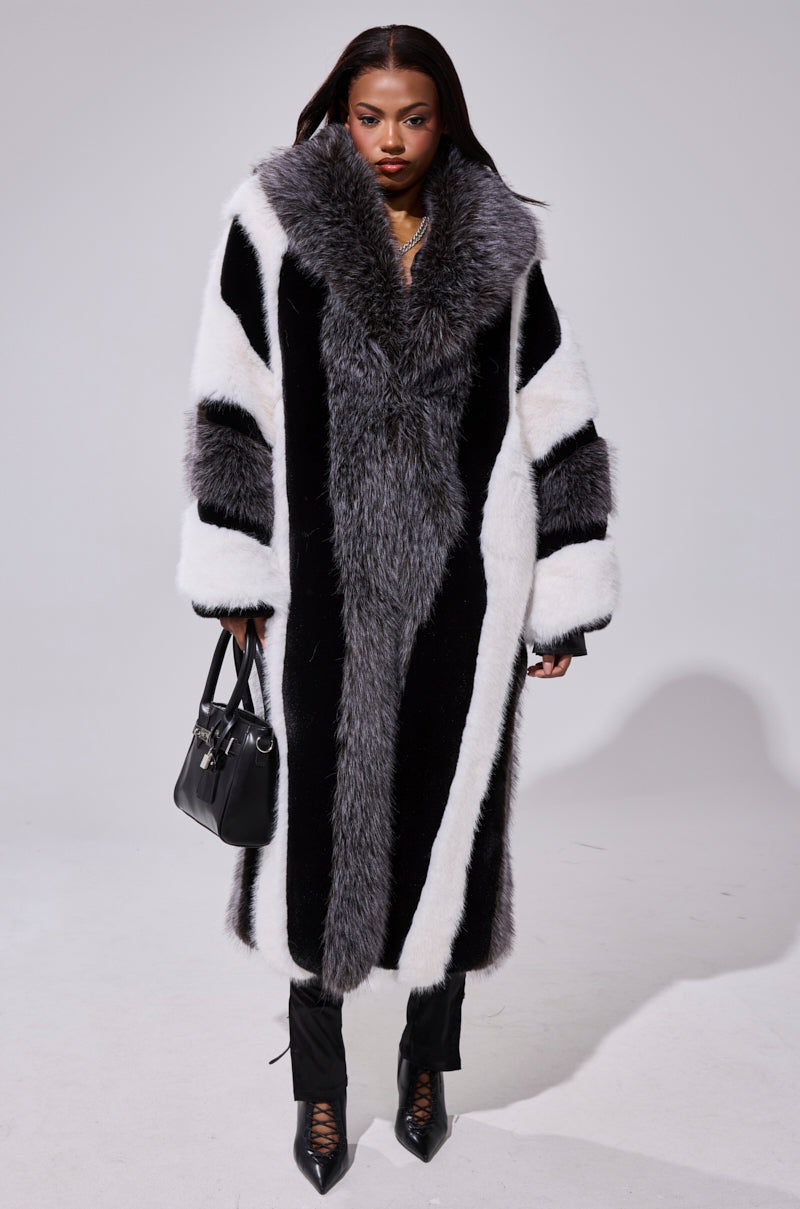 A woman stands facing forward in the WANT IT ALL FAUX FUR longline coat with black, white, and gray tones. She carries a black handbag and wears black pants and pointed-toe shoes against a plain, light-colored backdrop.