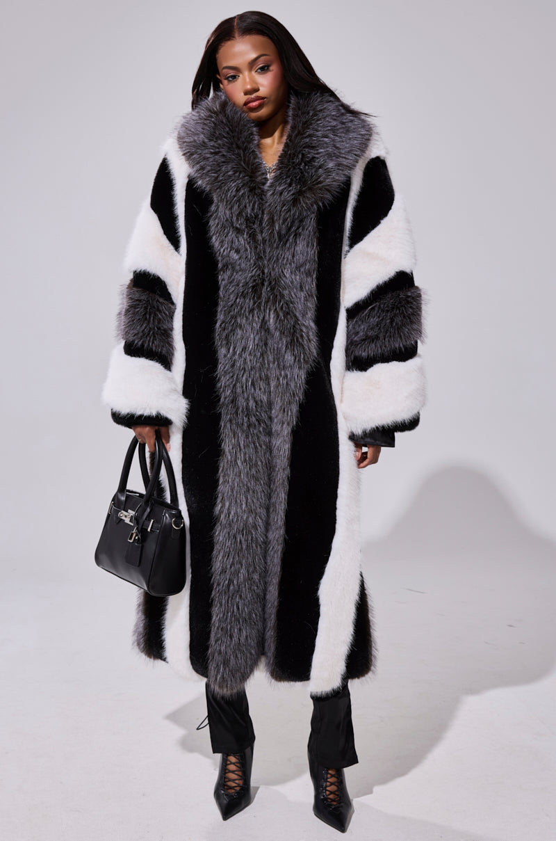 Wearing the WANT IT ALL FAUX FUR longline color block coat with wide gray trim, black pants, lace-up heeled boots, and carrying a black handbag, a person stands against a plain background.