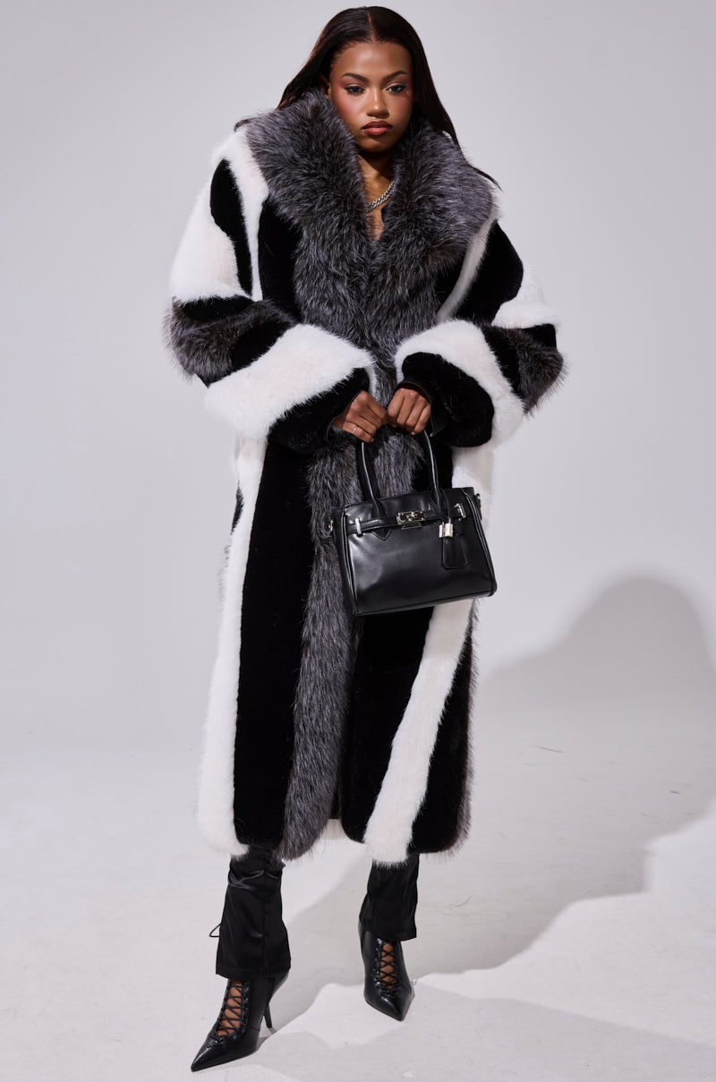 A woman poses in the WANT IT ALL FAUX FUR coat, featuring a longline black and white silhouette with a large gray collar, paired with black lace-up boots and a black handbag against a plain light background.