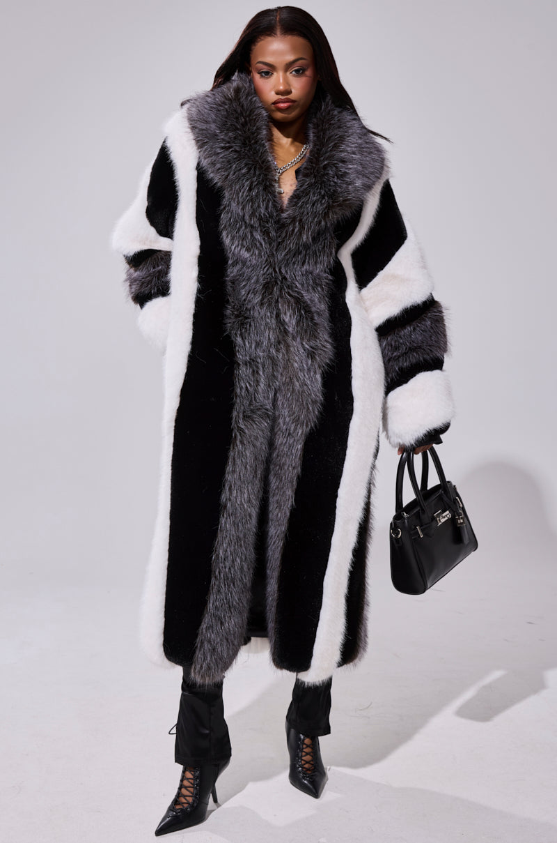 A woman stands confidently in the WANT IT ALL FAUX FUR coat with a longline silhouette, paired with black pants, lace-up heeled boots, and a black handbag against a plain light background.