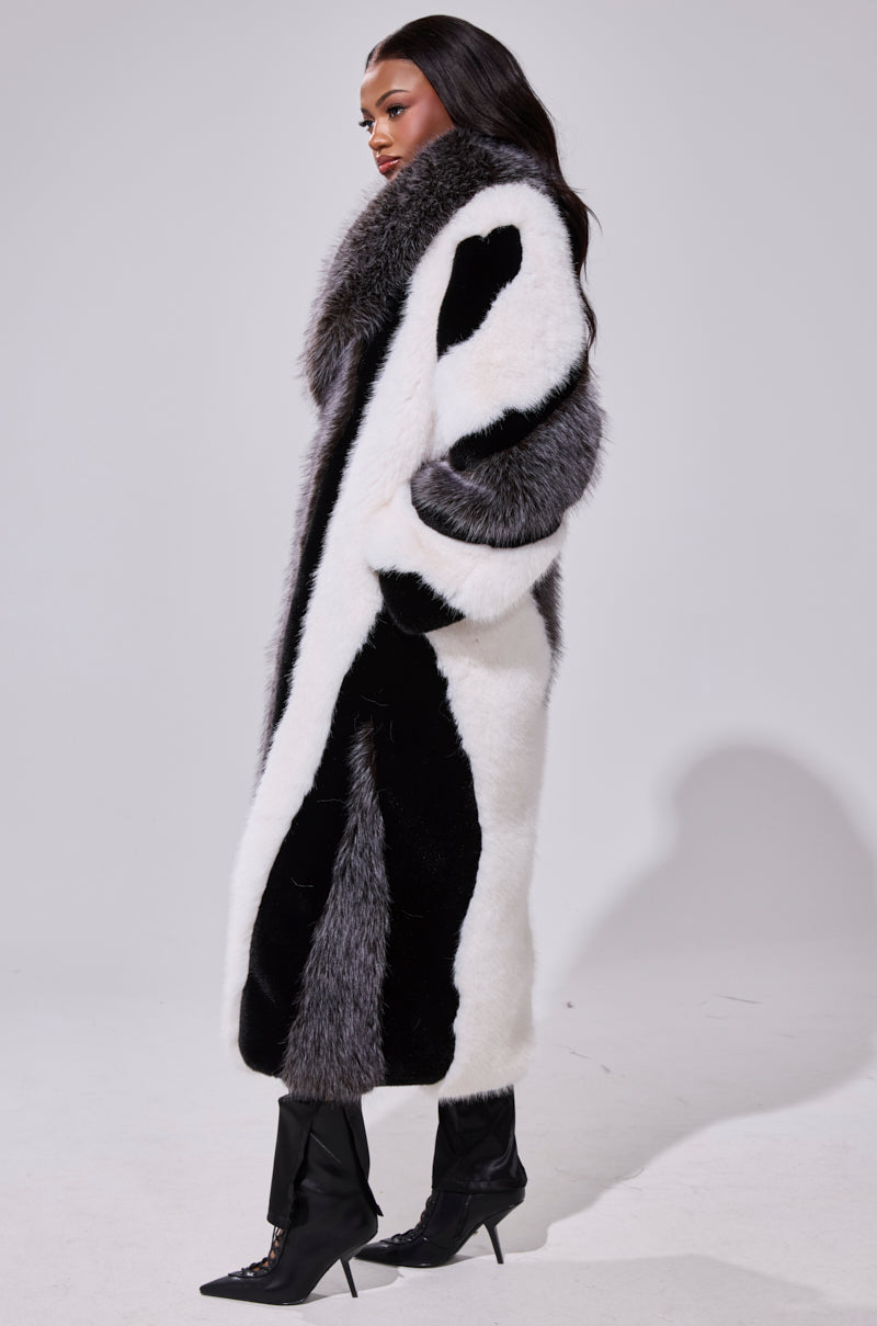 A woman stands in profile wearing the WANT IT ALL FAUX FUR coat, featuring black, white, and gray color blocks in a longline silhouette. She pairs it with black heeled boots against a plain light gray background.