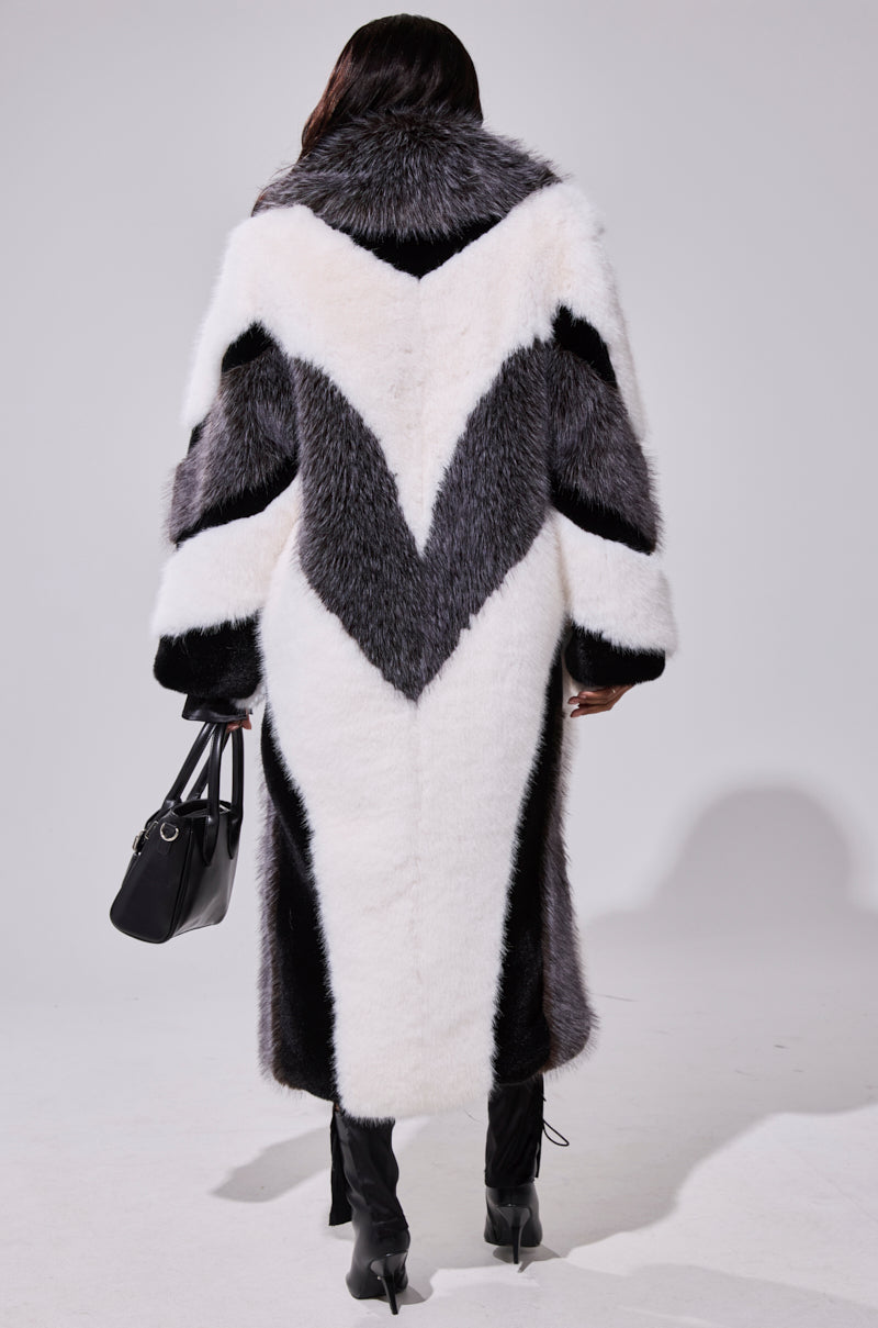 A person is seen from behind wearing the WANT IT ALL FAUX FUR longline coat with bold black, white, and gray chevron patterns, paired with a black handbag and black heeled boots.