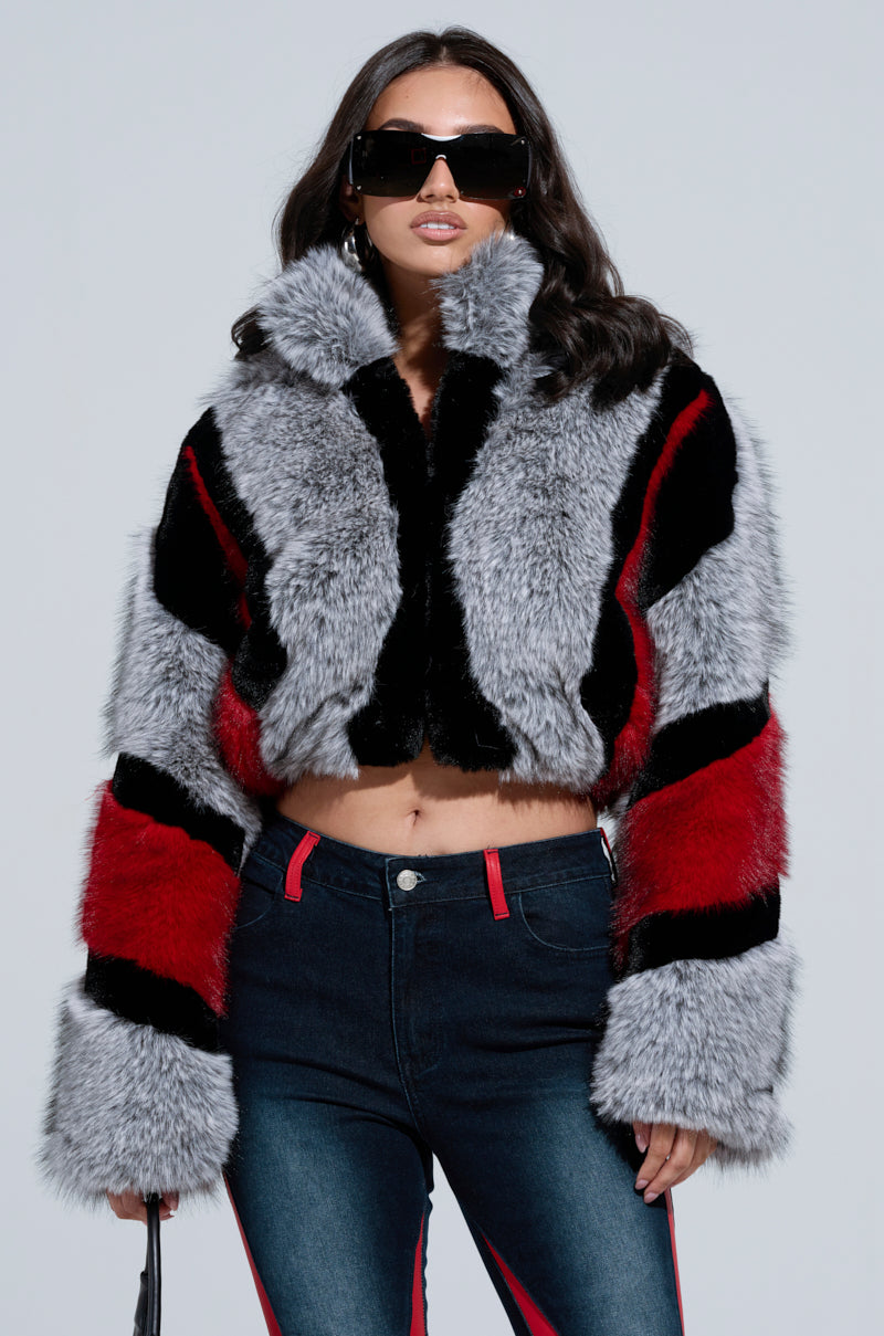 A person wearing the stylish TORONTO FAUX FUR COAT, featuring a chic mix of black, red, and gray patterns, is standing with their hands in their pockets. They have long dark hair and are sporting large black sunglasses along with dark jeans accented with red detailing.