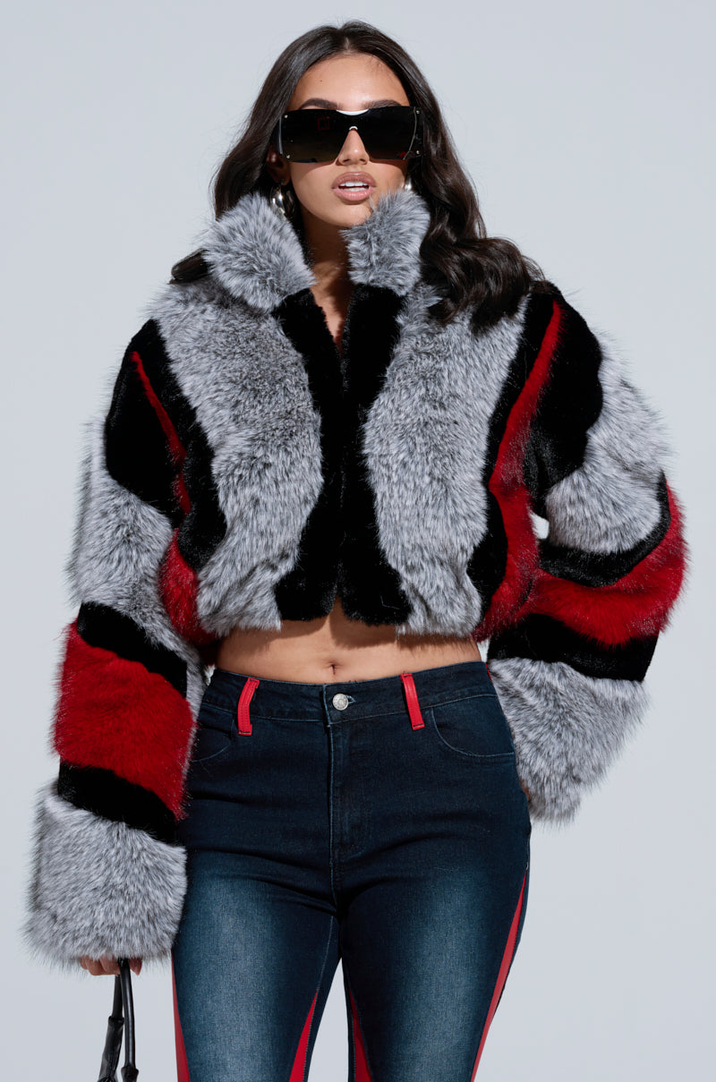 A woman confidently stands wearing the TORONTO FAUX FUR COAT, showcasing its chic color block pattern of black, red, and gray. Her style is further enhanced by dark sunglasses and form-fitting jeans accented in red against a simple background.