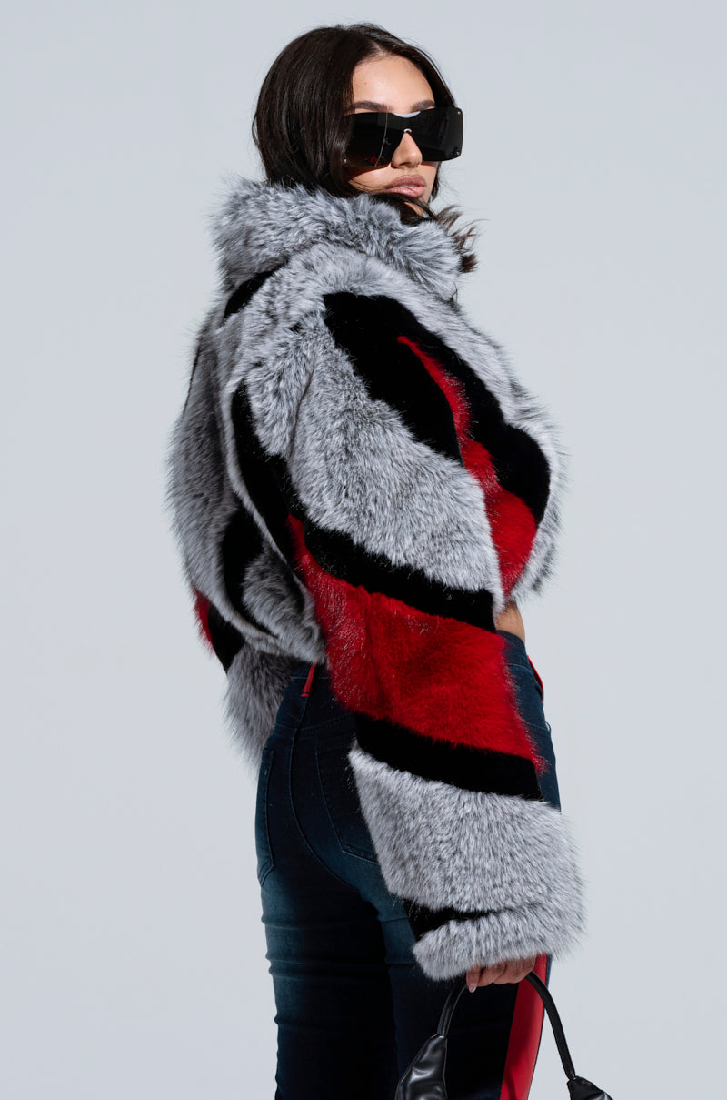 A woman shows off the TORONTO FAUX FUR COAT, which features a stylish gray and red striped color block pattern. She pairs it with jeans and accessorizes with large black sunglasses while her long dark hair flows freely. Holding a black bag, she faces backward with her head slightly turned toward the camera.
