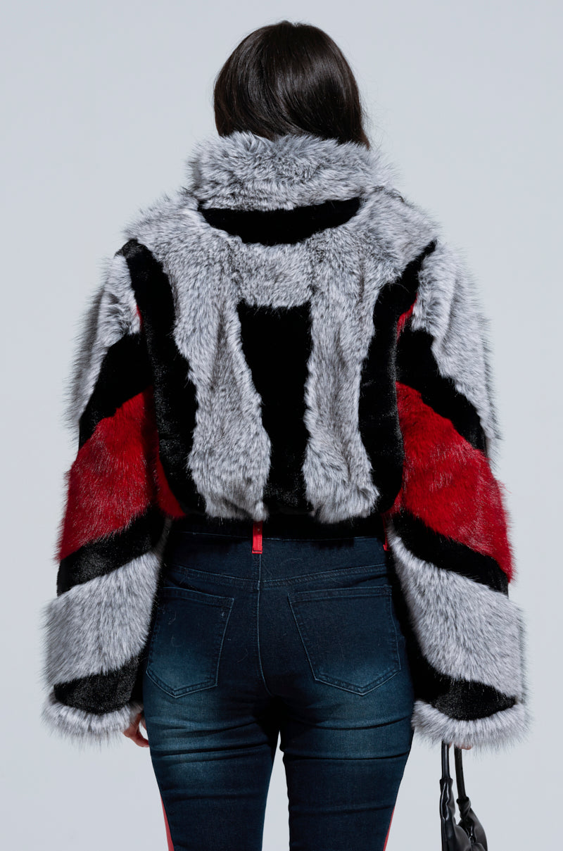 A rear view of a person donning the TORONTO FAUX FUR COAT, featuring a cropped silhouette and multicolored color block patterns in red, black, and gray. They have dark hair and carry a black handbag that pairs well with their dark jeans.