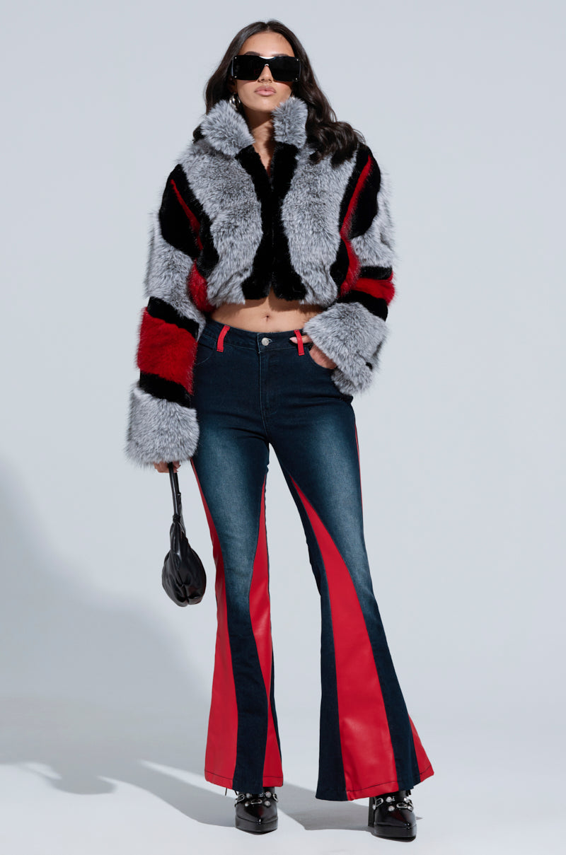 A person wearing large sunglasses and the TORONTO FAUX FUR COAT, which features a color block pattern of black, red, and gray stripes, stands confidently in flared jeans with red panels. They hold a small black handbag, presenting a striking silhouette against the plain backdrop.