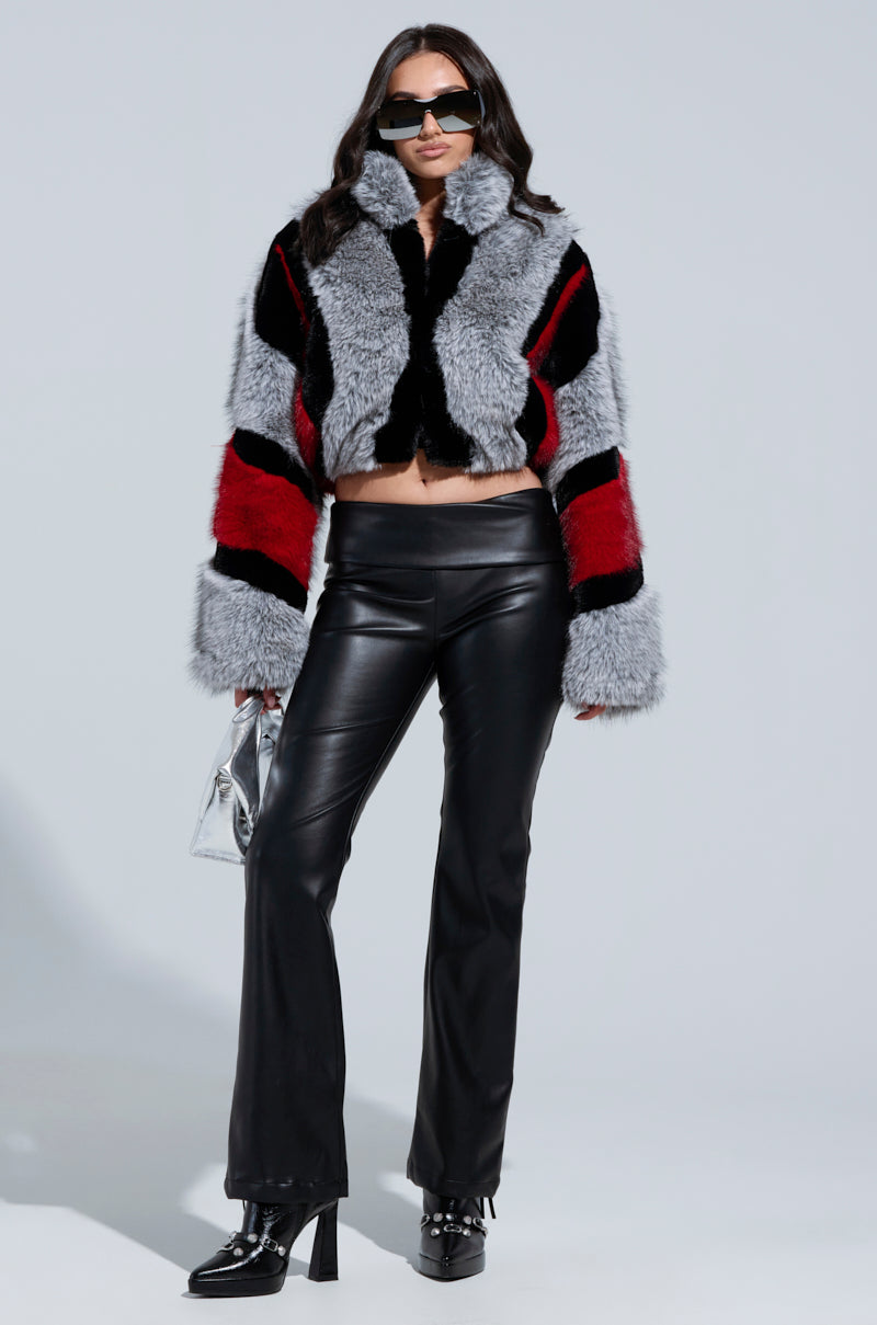 A person is wearing the stylish TORONTO FAUX FUR COAT, featuring a color block pattern of black, red, and gray patches. The coat is paired with sleek black leather pants and glossy high-heeled boots. They are accessorized with a small metallic bag and large sunglasses while standing against a plain background.
