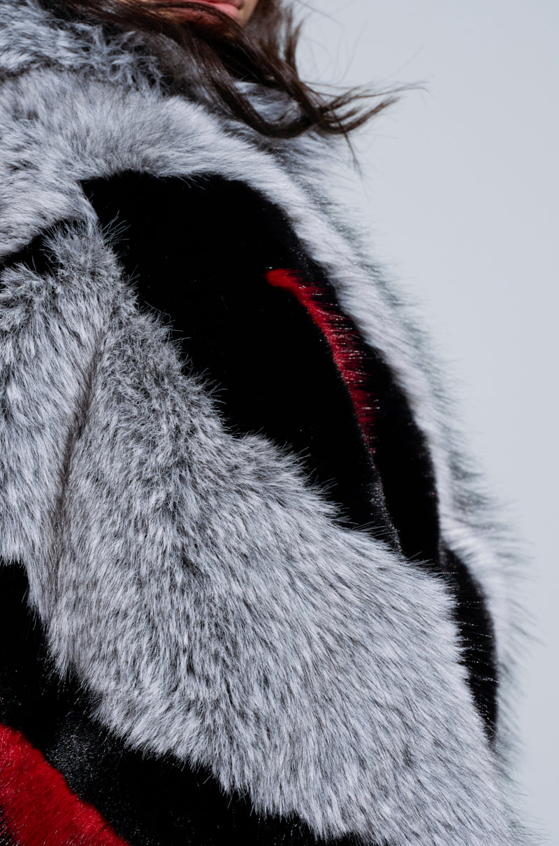 Close-up of the TORONTO FAUX FUR COAT, showcasing a blend of gray, black, and red patterns. The texture looks soft and plush, with only the cropped silhouette of the wearer visible against a neutral background, adding an intriguing contrast.
