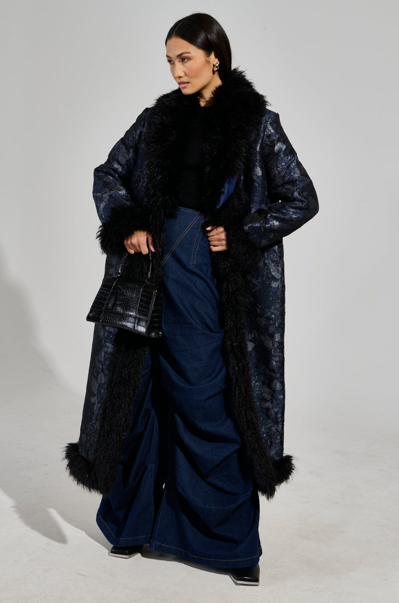 A woman in an opulent, dark WILD CARD BROCADE TRENCH COAT featuring a mohair collar stands against a plain background, wearing wide-leg denim pants and a black top. She carries a textured black handbag and elegant earrings dangle from her ears.