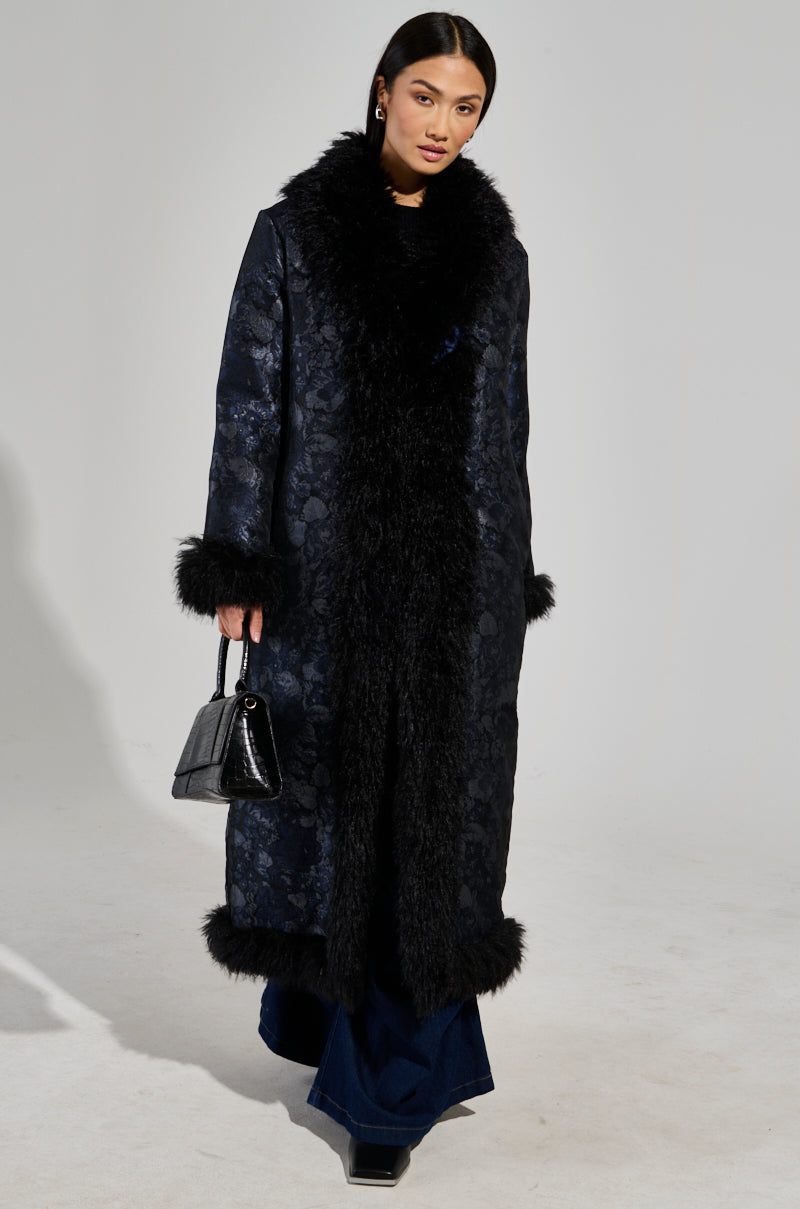 A person stands against a plain background, wearing the Wild Card Brocade Trench Coat with opulent fur accents on the mohair collar, cuffs, and hem. They carry a small black handbag, completing the ensemble with dark pants and shoes.