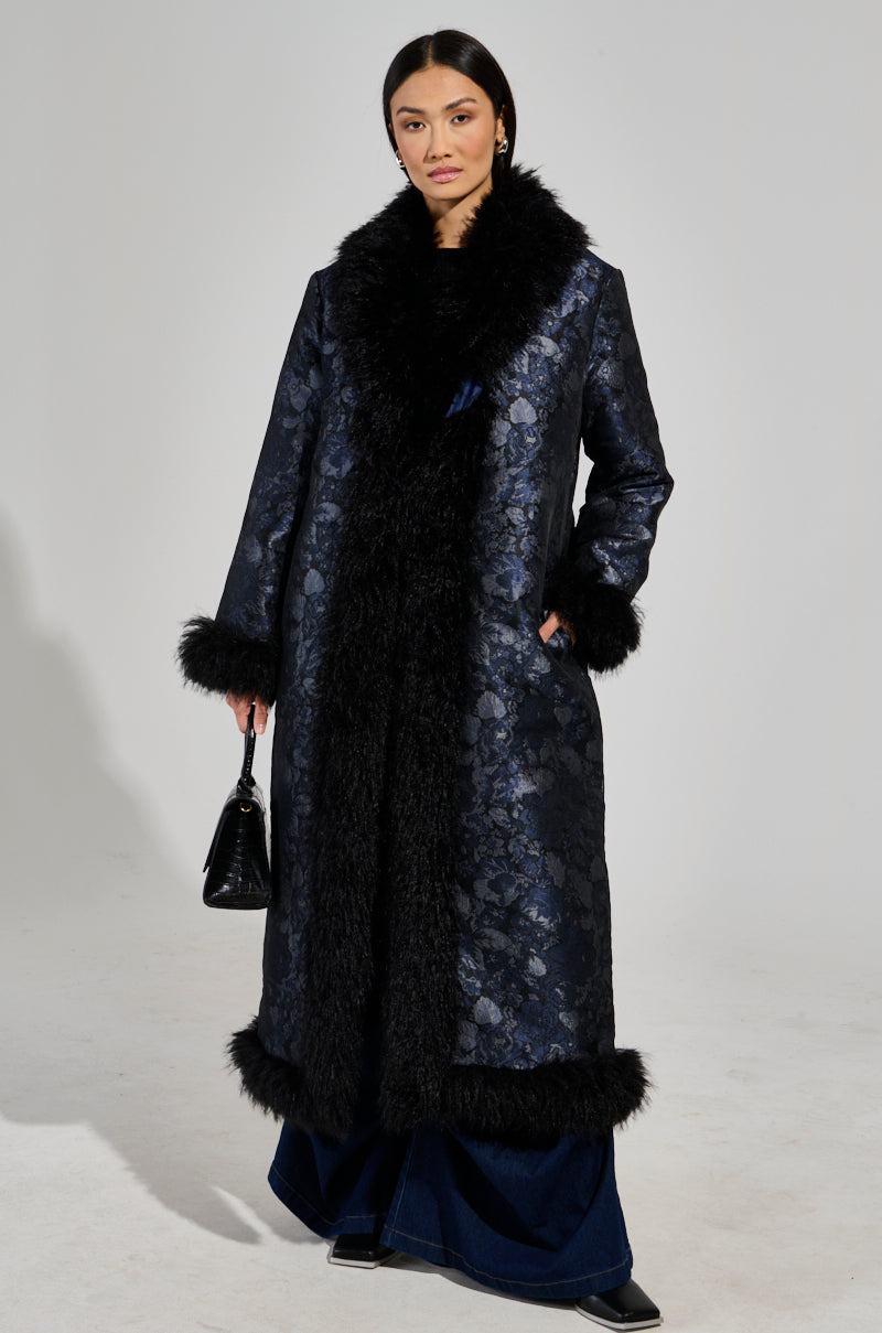 A person confidently stands wearing the WILD CARD BROCADE TRENCH COAT, which features a long, dark brocade design complemented by black, fluffy mohair trim on the collar, cuffs, and hem. They hold a small black handbag against a plain, light-colored background.