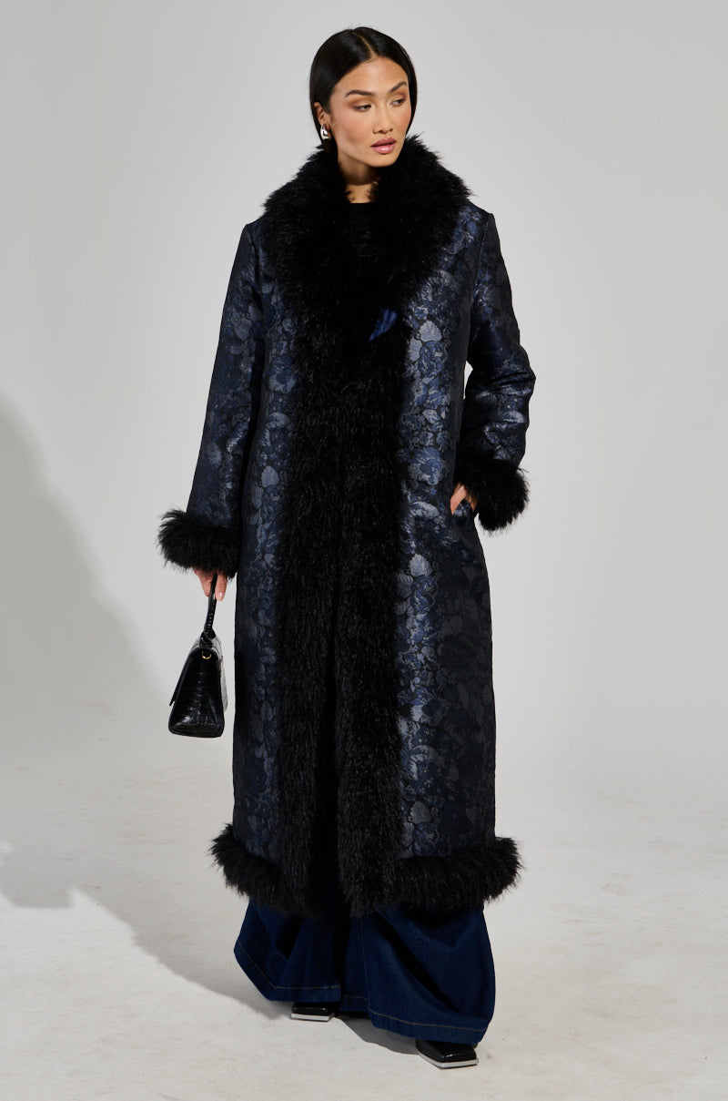 A woman stands wearing the WILD CARD BROCADE TRENCH COAT, enhanced with a baroque floral pattern and featuring a mohair collar and cuffs. She holds a small black handbag, her hair parted down the middle, and dark wide-legged pants peeking from beneath complete her elegant ensemble.