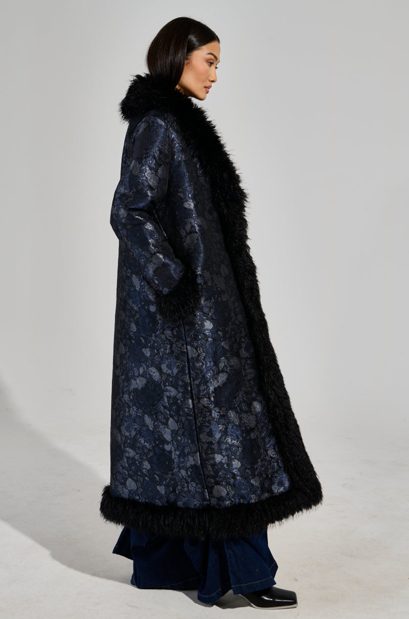 A person is wearing the WILD CARD BROCADE TRENCH COAT, designed with a mohair collar and adorned in a dark floral baroque pattern, complete with fur trim along the cuffs and hem. They pair it with wide-leg dark blue pants and black shoes against a plain, light-colored background.