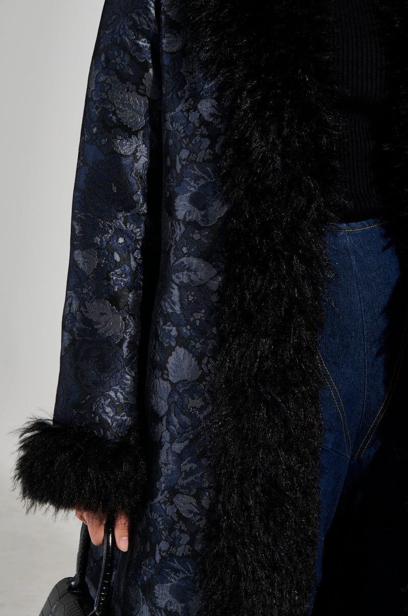 A person wearing the WILD CARD BROCADE TRENCH COAT, featuring an elegant baroque floral pattern and a mohair collar, holds a black woven handbag. They are also dressed in dark denim jeans, set against a plain light background.