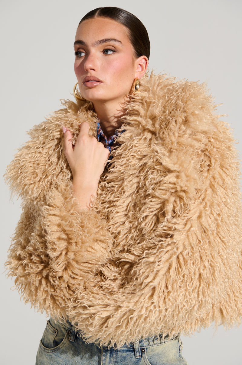 A person in a JOLENE FAUX FUR COAT, featuring a large, textured beige design, pairs it with blue jeans while posing against a neutral background. Their hair is pulled back, highlighting gold hoop earrings that glint as they gaze pensively to the side—prepared for a winter's night out.