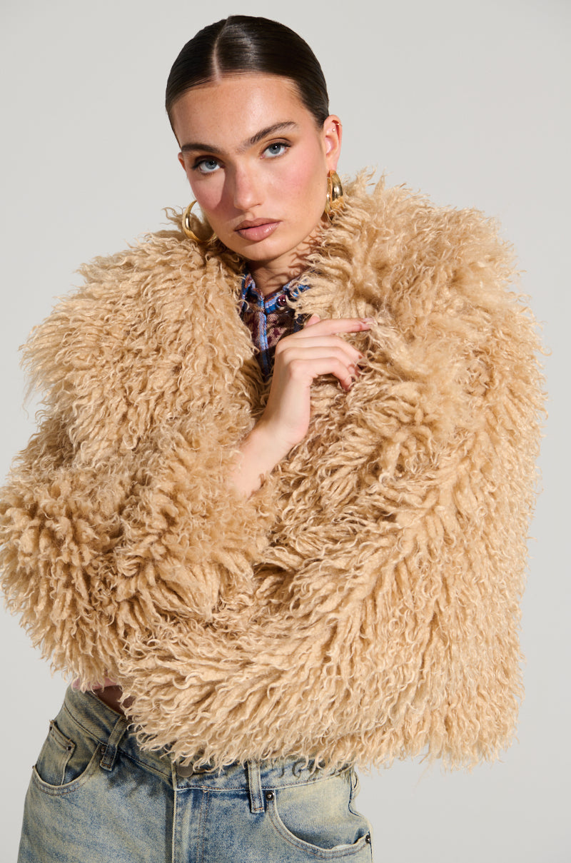 Wrapped in the JOLENE FAUX FUR COAT, a plush beige masterpiece ideal for a chilly evening out, they pose with confidence. Their sleek hairstyle and oversized hoop earrings harmonize beautifully with light blue jeans. The coat's cropped design contrasts strikingly against the neutral backdrop, highlighting its bold texture.