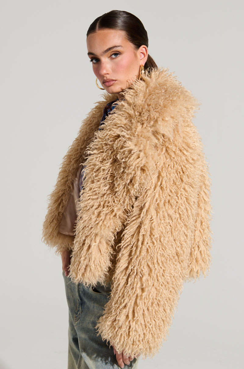 Against a plain backdrop, a person exudes confidence in the JOLENE FAUX FUR COAT, its shaggy beige texture draped over a light ensemble paired with jeans. Their hair slicked back and cropped silhouette perfectly capture the essence of Winter's Night Out as they gaze thoughtfully into the distance.