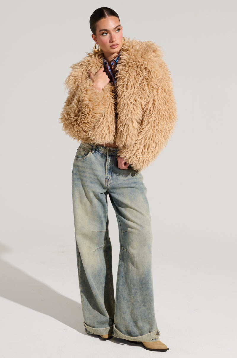 Dressed in the stylish JOLENE FAUX FUR COAT and wide-leg jeans, a person confidently poses against a light gray backdrop. Their slicked-back hair and boots perfectly complement the ensemble, ideal for a winter's night out.