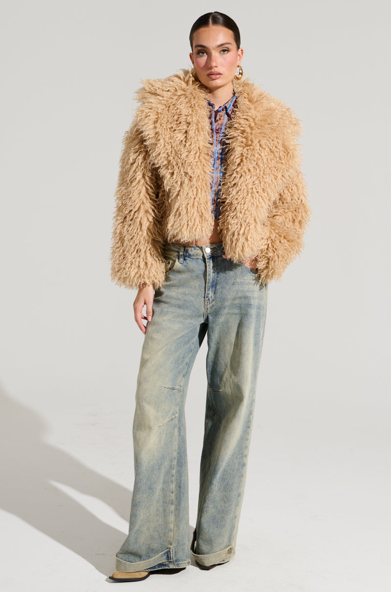 A person stands confidently in a cropped silhouette, featuring the JOLENE faux fur coat, blue wide-legged jeans, and beige shoes. The plain background enhances the focus on this stylish ensemble, perfect for a winter's night out.