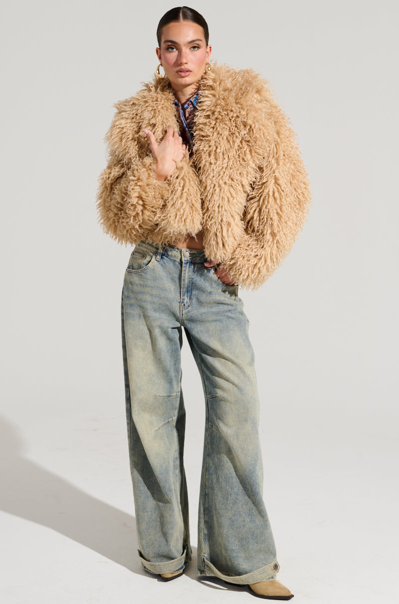 A person stands confidently in a cropped silhouette, showcasing the JOLENE FAUX FUR COAT and wide-leg jeans. With their hair pulled back and one hand on their waist, they project a stylish and modern vibe against the plain background, perfect for a winter's night out.