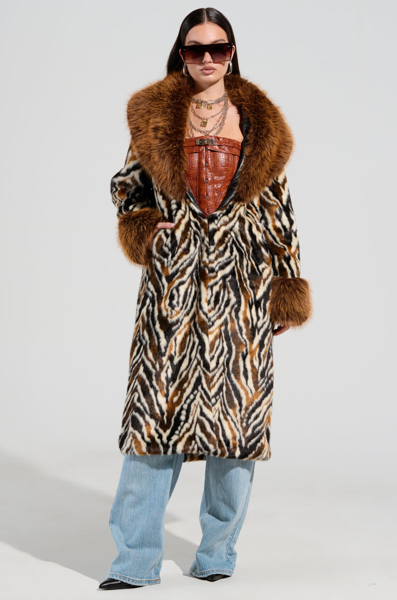 A person wearing sunglasses is styled in an ALANA FUR COAT, a zebra-inspired faux fur maxi coat featuring a bold brown collar and cuffs. They complement it with a brown top and wide-legged blue jeans, against a plain background.