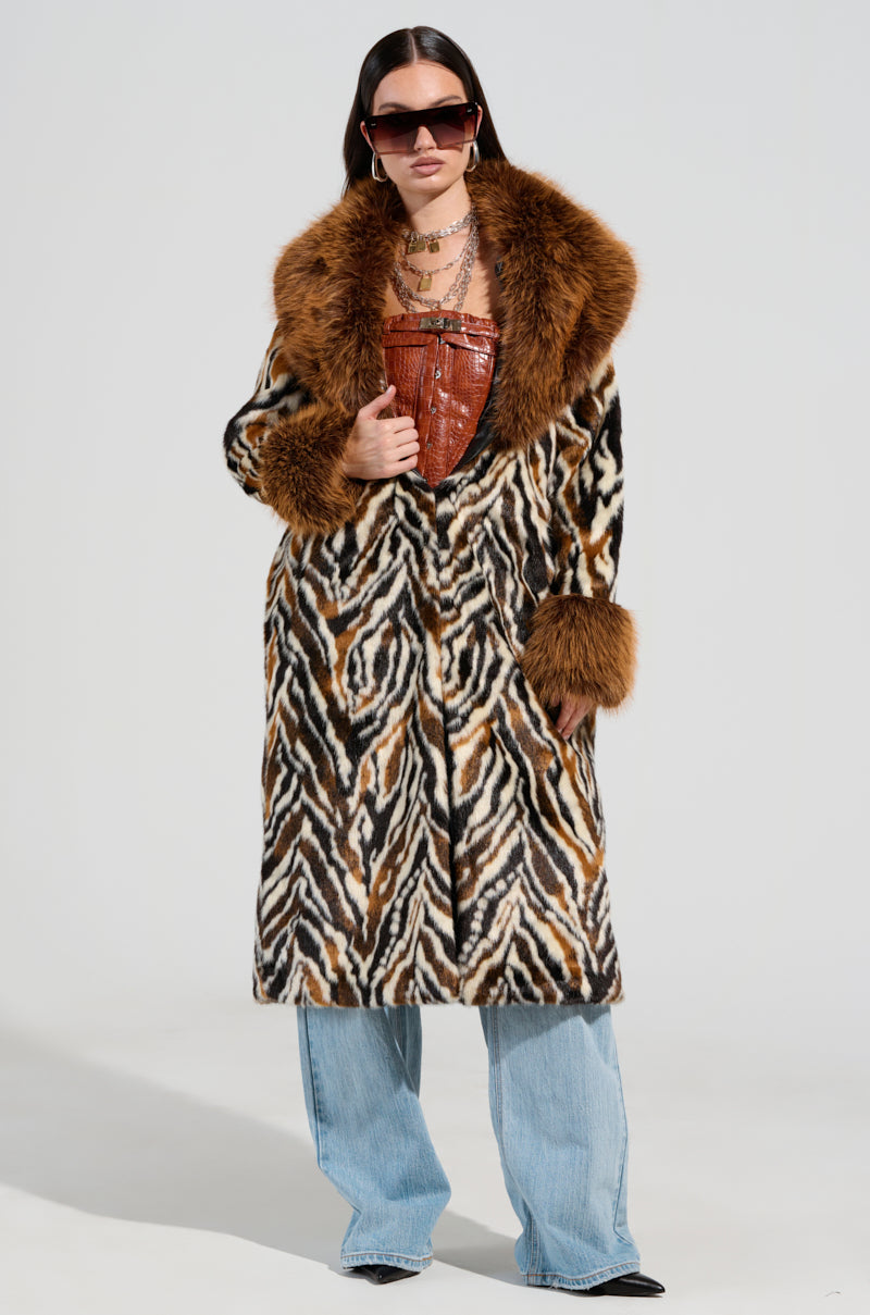 A person confidently stands wearing the ALANA FUR COAT, a maxi-length coat with a zebra-inspired pattern and brown fur trim. They hold a brown leather bag, rock oversized sunglasses, and sport wide-legged light blue jeans against a plain, light-colored background.