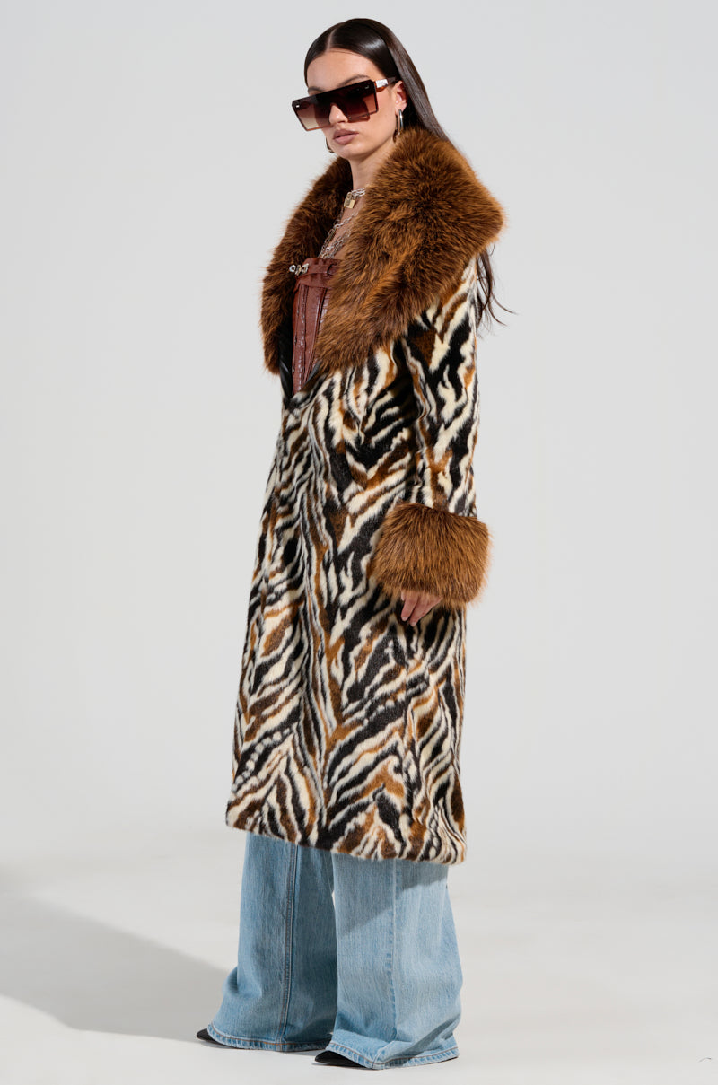 A person wearing large dark sunglasses stands against a plain background, donning the stunning ALANA FUR COAT, featuring zebra-inspired patterns and a faux fur collar. The coat is elegantly paired with wide-leg blue jeans, highlighting a stylish blend of black, white, and brown stripes.