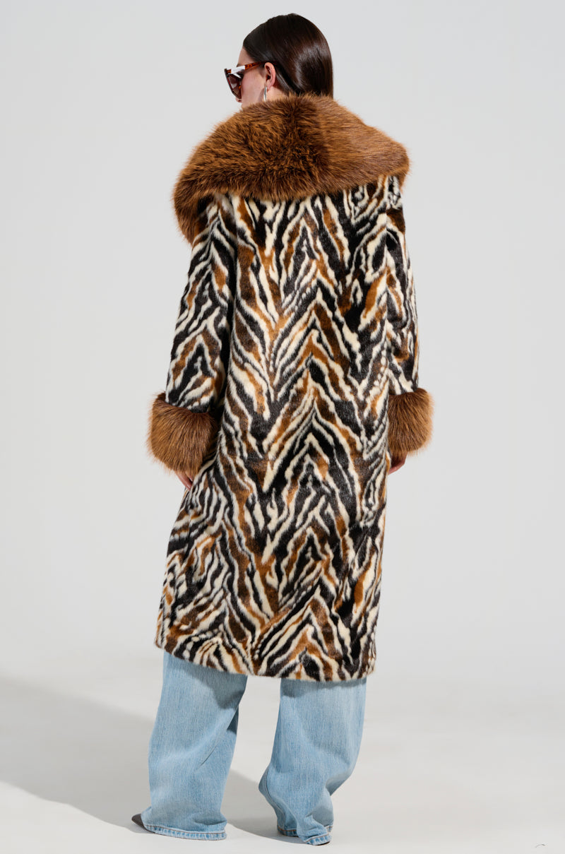 Wearing an ALANA FUR COAT with a striking zebra-inspired pattern and brown cuffs, a person is seen from behind. They pair it with light blue jeans, stylish sunglasses, and have long dark hair. The plain gray backdrop highlights their daring fashion statement.
