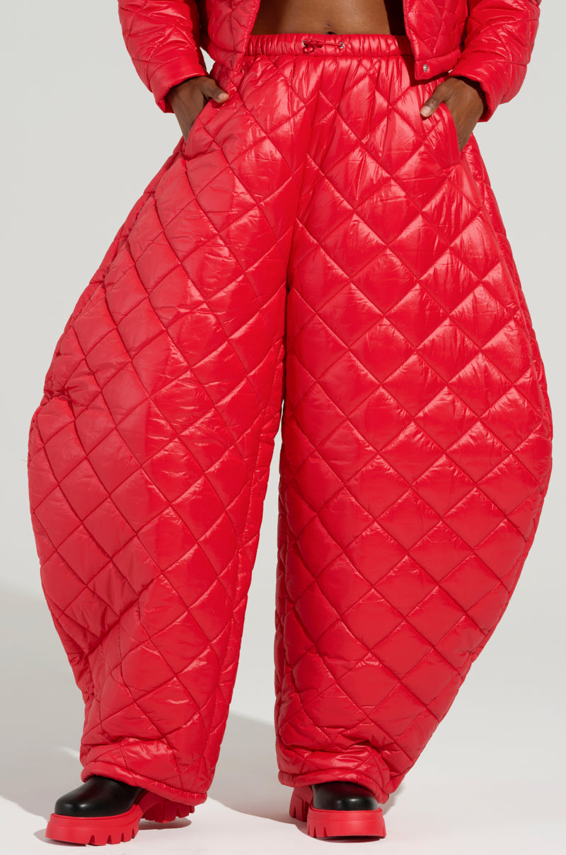 A person wearing the AKIRA Label VIVI Oversized Puffer Pants in Red stands with hands in pockets. The dynamic pants, paired with black and red boots, create a striking contrast against the plain background. The image artfully highlights this bold lower-body ensemble.