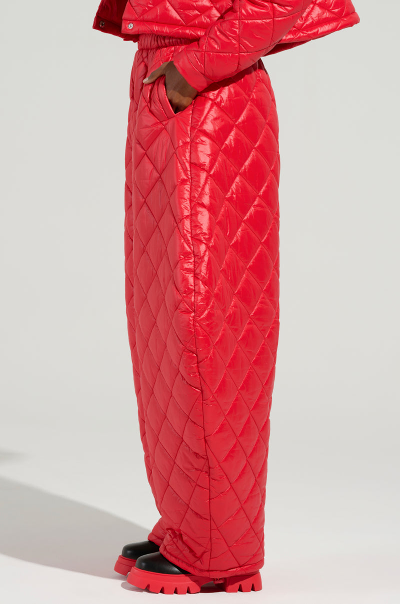 A person wearing the striking VIVI OVERSIZED PUFFER PANTS IN RED from AKIRA Label, featuring a bright red quilted design with a high waistband and side pockets. The glossy pants are paired with chunky black boots that have thick red soles, creating a bold look against a plain background.