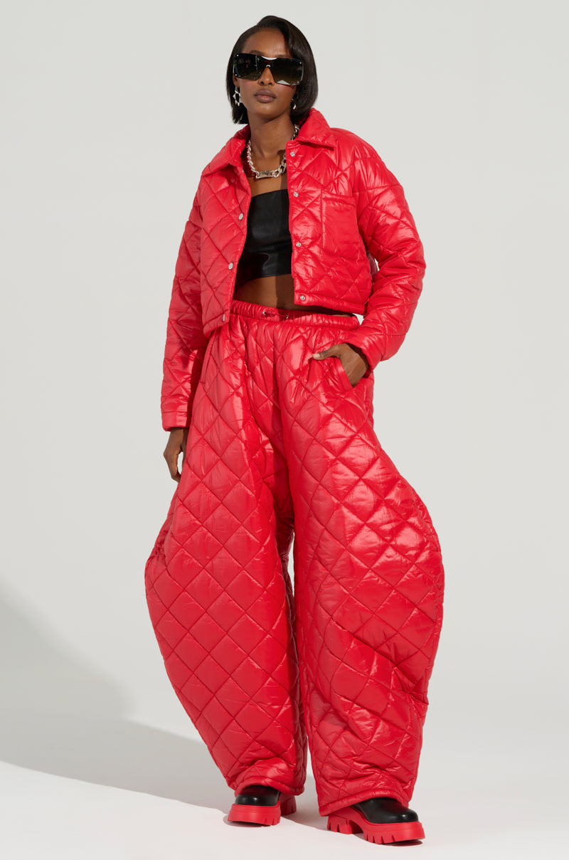 Dressed in the VIVI OVERSIZED PUFFER PANTS IN RED from AKIRA Label and a coordinating red jacket over a black top, the individual adds flair with large sunglasses and chunky red and black shoes, creating a bold and fashion-forward ensemble.