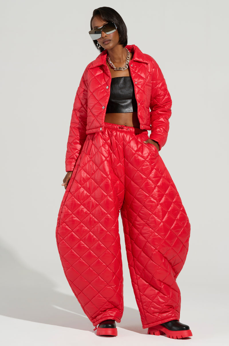 A person dressed in a vibrant red quilted jacket and Vivi Oversized Puffer Pants in Red, paired with a black crop top and chunky red and black boots. They complete their look with futuristic sunglasses and a silver chain necklace as they stand confidently against a plain background.