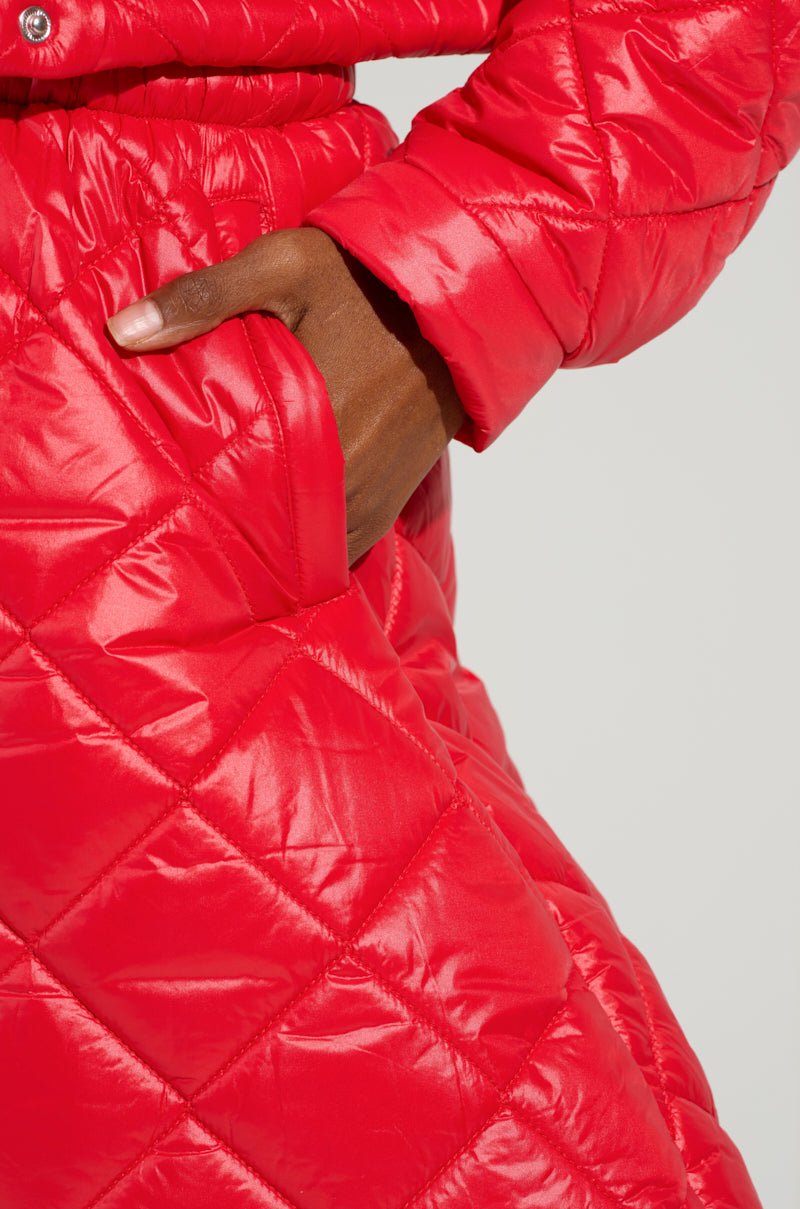 A close-up of a person wearing a red quilted jacket from the AKIRA Label shows their right hand in a pocket. The jacket's diamond-shaped pattern perfectly complements their bold and stylish VIVI Oversized Puffer Pants in red.