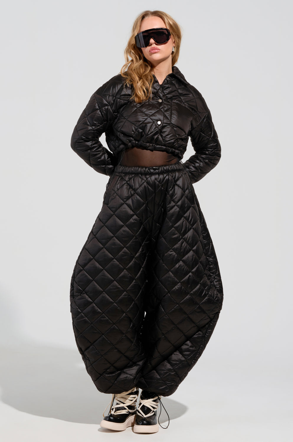 A person poses confidently in matching black quilted attire, showcasing the AKIRA Label Vivi collection’s oversized puffer pants and a coordinating cropped jacket. They sport large sunglasses with their hands casually tucked into the pockets of their VIVI Oversized Puffer Pants in Black.