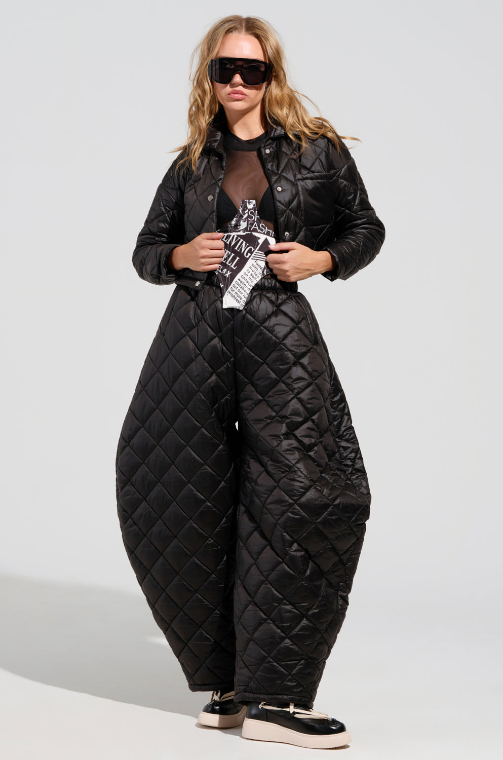 A person wearing the VIVI Oversized Puffer Pants in black, reminiscent of the AKIRA Label Vivi style, along with a matching jacket styled with sunglasses and black shoes, stands against a plain background.