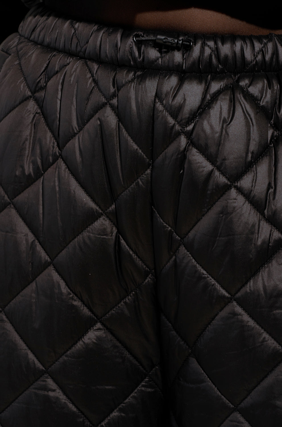 Close-up of the black Vivi Oversized Puffer Pants in a quilted design, showcasing a shiny finish. The fabric features a diamond-shaped pattern accentuated by lighting. It has an elastic waistband with a discreet front drawstring, all against a dark backdrop.