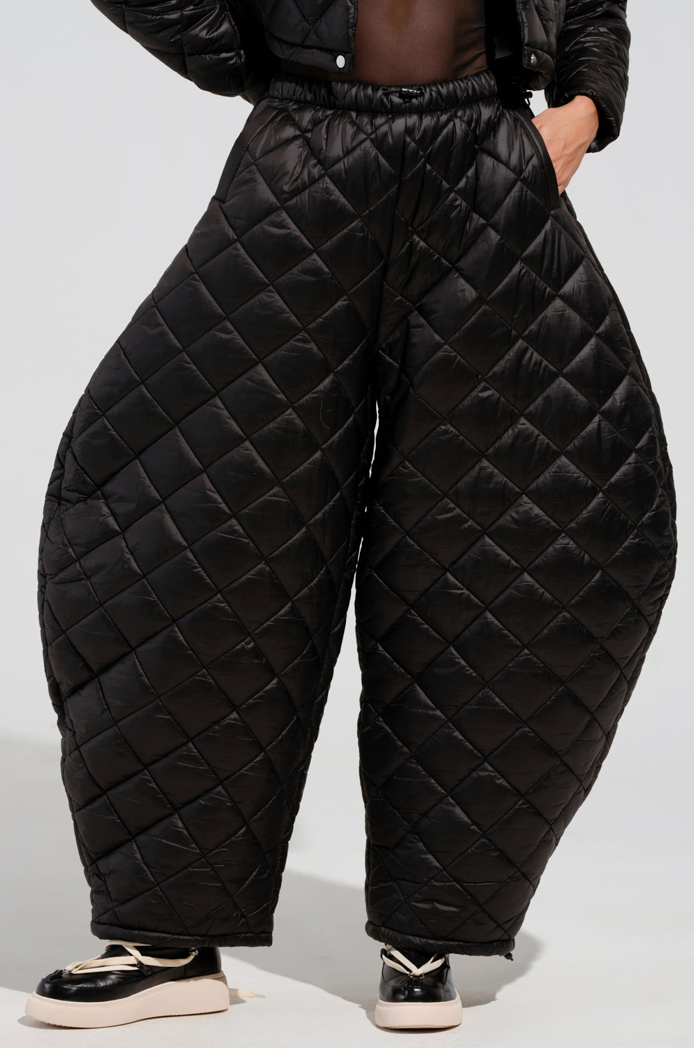 A person wearing the VIVI Oversized Puffer Pants in Black with a striking diamond pattern. The unique, wide silhouette of these black pants pairs effortlessly with sleek black and white sneakers. The simple, light-colored backdrop enhances the boldness of the outfit.