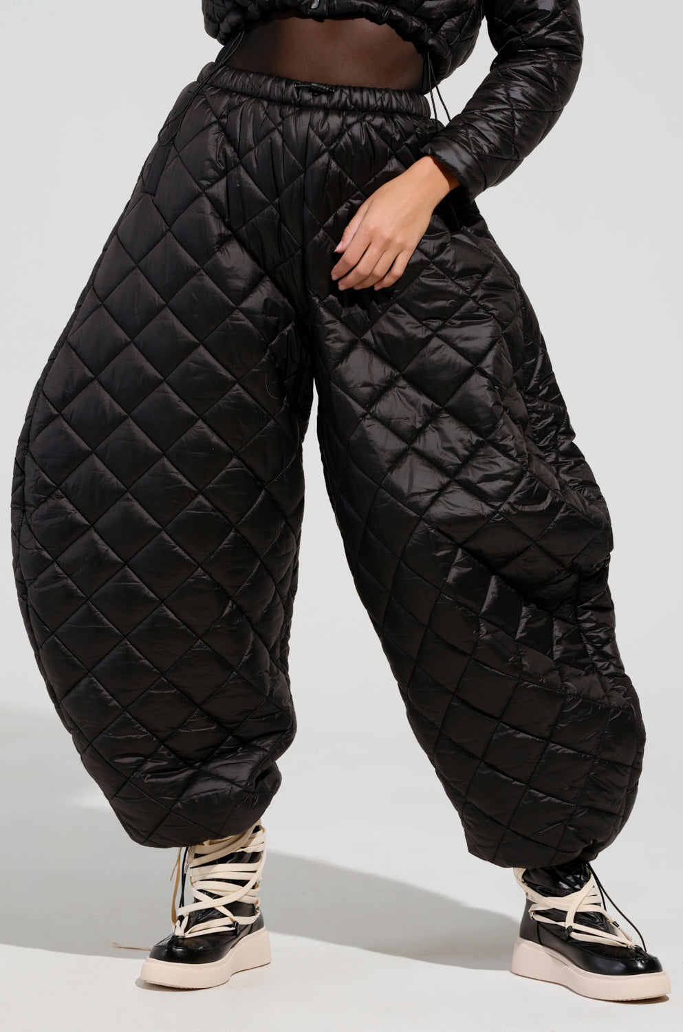 Someone is wearing the VIVI OVERSIZED PUFFER PANTS IN BLACK by AKIRA Label, paired with high-top sneakers that feature white laces and tan soles. The plain, light-colored background perfectly complements the striking outfit.