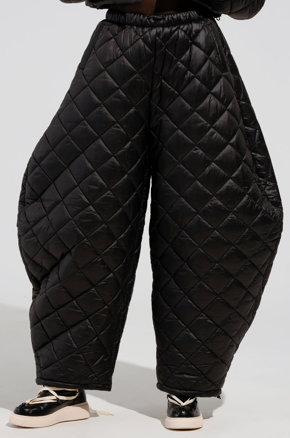 An individual is sporting the VIVI Oversized Puffer Pants in Black from AKIRA Label, featuring a quilted diamond pattern and a puffy, exaggerated design. They're paired with shiny black and white shoes against a simple, light-colored background.