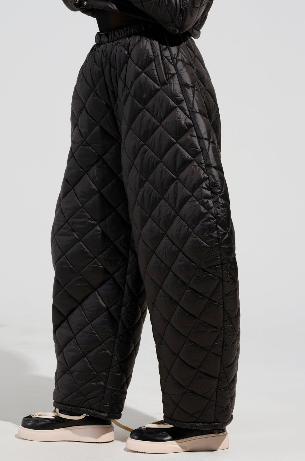A person is standing against a plain background wearing the VIVI Oversized Puffer Pants in Black from AKIRA Label, paired with stylish black-and-white platform shoes. The pants are characterized by their diamond pattern and a comfortable elastic waistband.