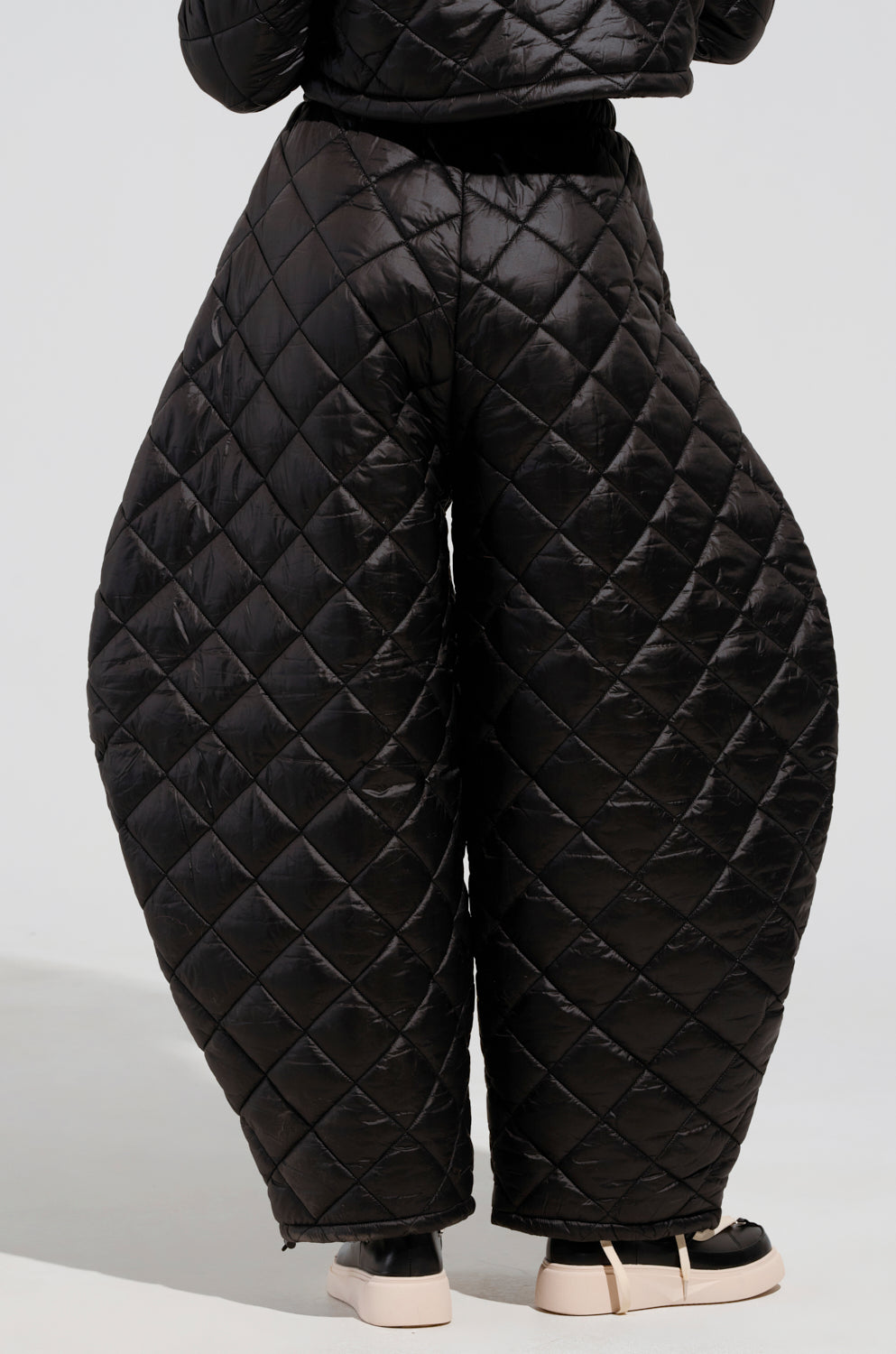 A person wearing the VIVI OVERSIZED PUFFER PANTS IN BLACK, featuring a diamond quilted pattern and voluminous cocoon-like shape, stands on a light surface. The pants are effortlessly paired with black and white sneakers.