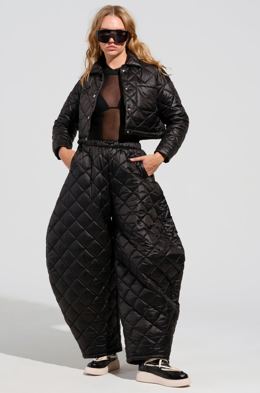 Wearing the VIVI Oversized Puffer Pants in black from AKIRA Label, paired with a matching cropped jacket, a sheer top, and a black bra. They complete the look with black sunglasses and chunky black-and-white shoes while standing confidently against a neutral background.